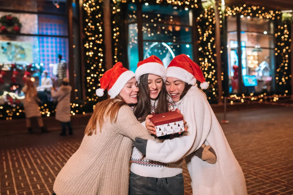 Tres amigas con gorros navideños se abrazan con alegría mientras sostienen un regalo, rodeadas de luces festivas en un ambiente decembrino.