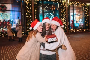 Tres amigas con gorros navideños se abrazan con alegría mientras sostienen un regalo, rodeadas de luces festivas en un ambiente decembrino.